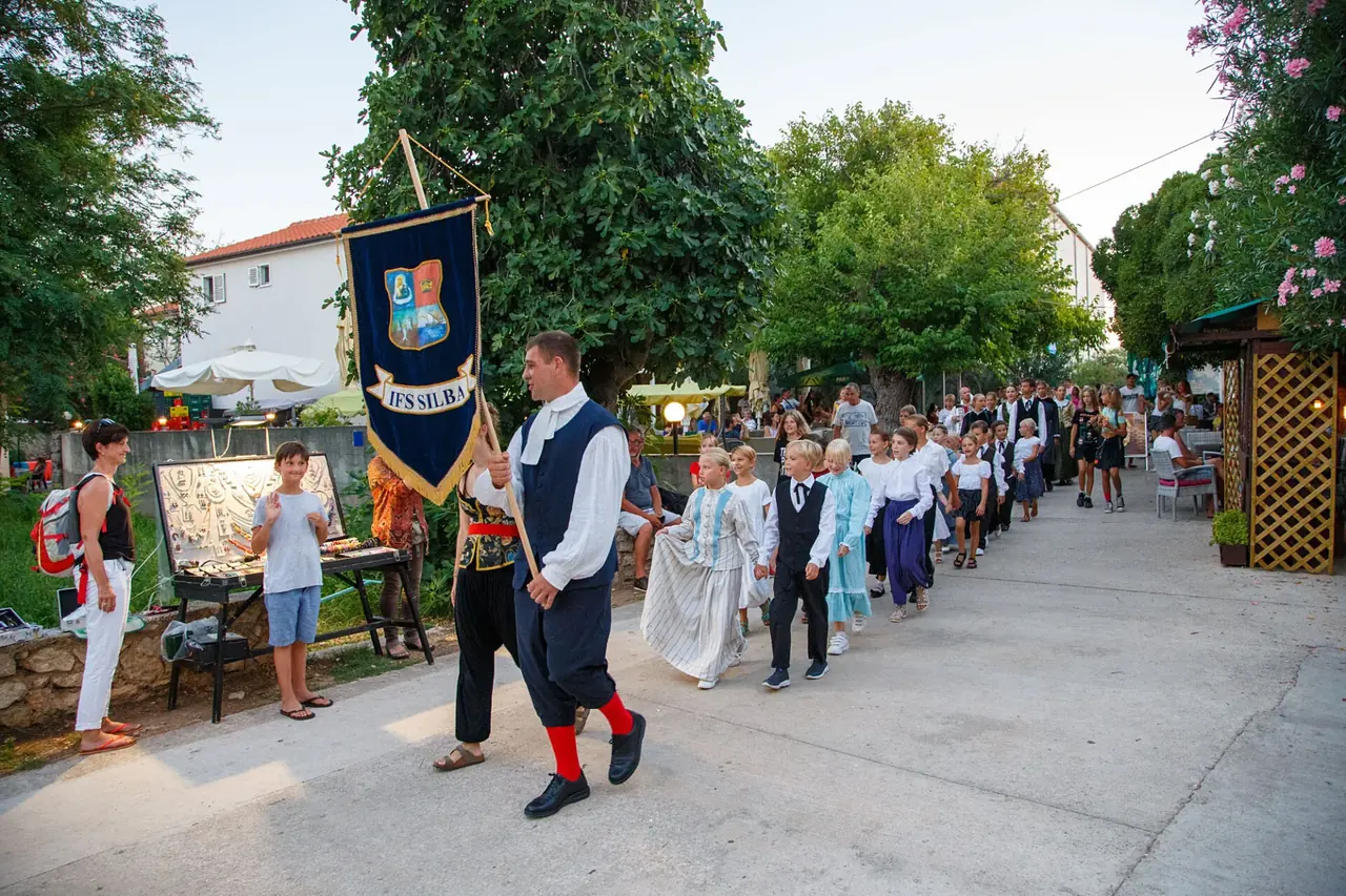 Đir po Silbi: plesač nosi barjak skupine na čelu povorke djece i odraslih u nošnjama kroz selo.